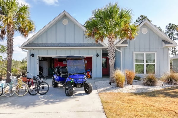 Modern light blue house with open garage containing blue golf cart, bicycles parked outside, palm trees, and sandy landscaping