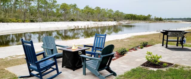 Waterfront patio with blue Adirondack chairs, fire pit table, and grill overlooking calm river and tree line